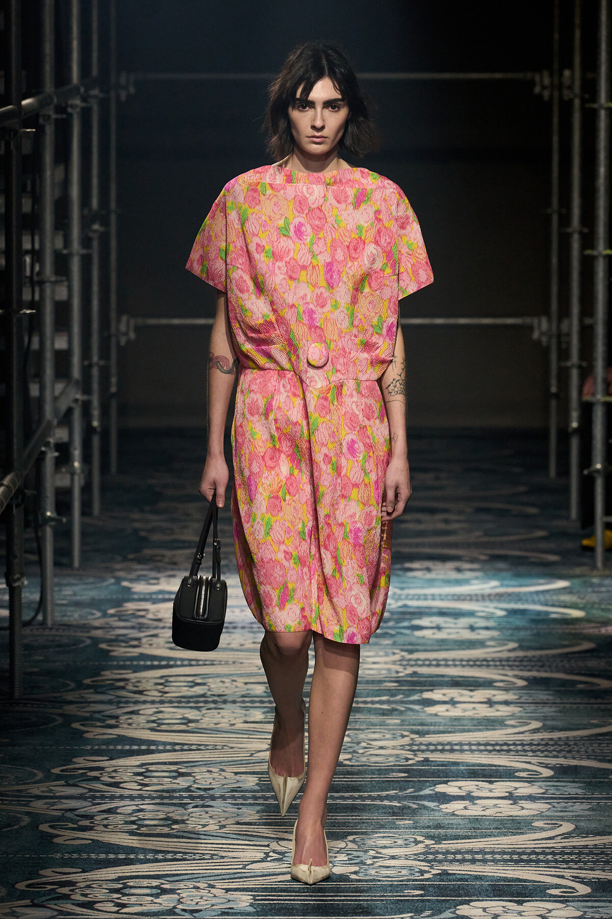 Model walking a fashion runway in a pink floral dress with a matching belt, carrying a small black handbag and wearing beige pointed heels.