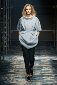 Model walking a runway in a light blue oversized knit top with large front pockets and rolled sleeves, paired with black trousers and black sandals.