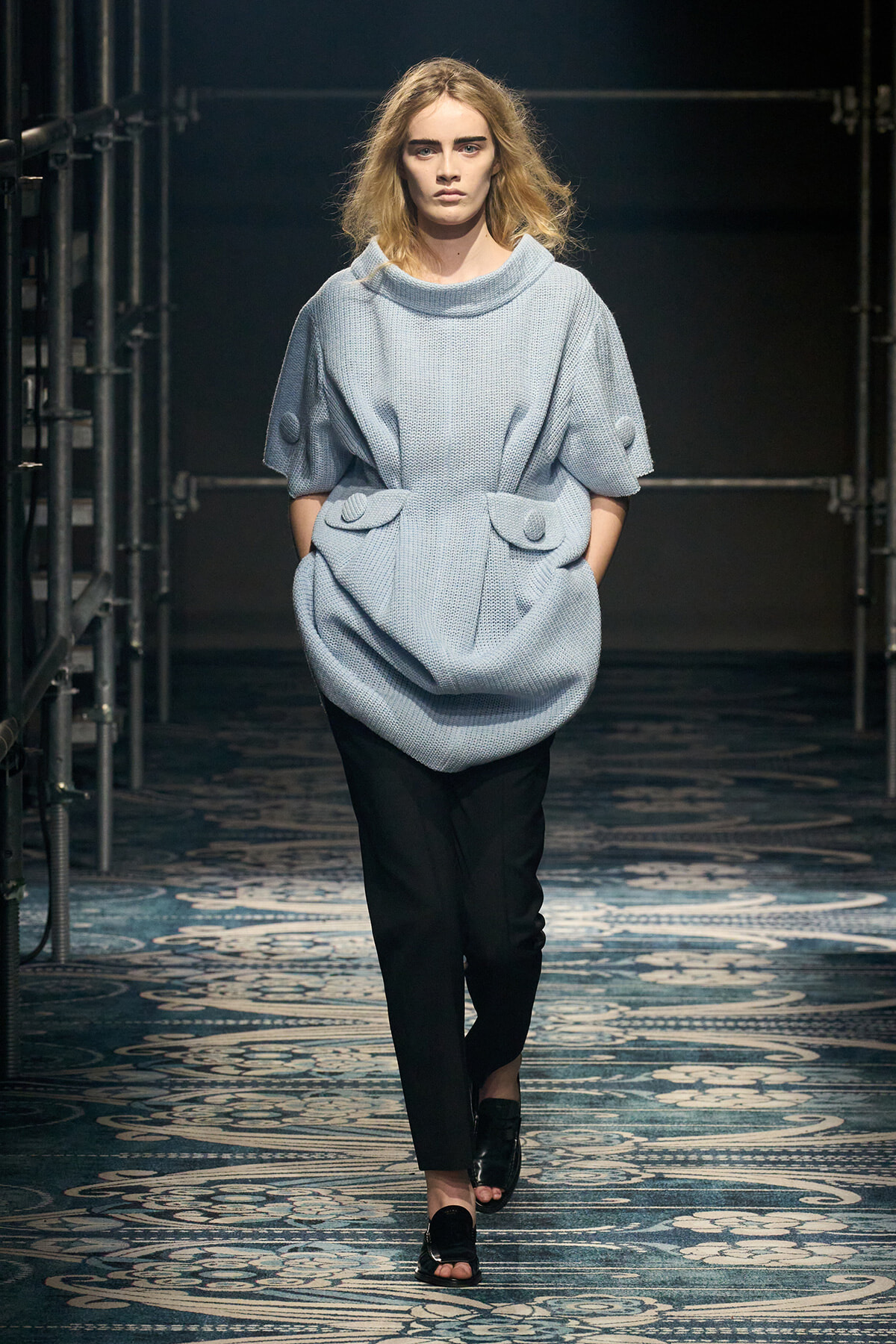 Model walking a runway in a light blue oversized knit top with large front pockets and rolled sleeves, paired with black trousers and black sandals.