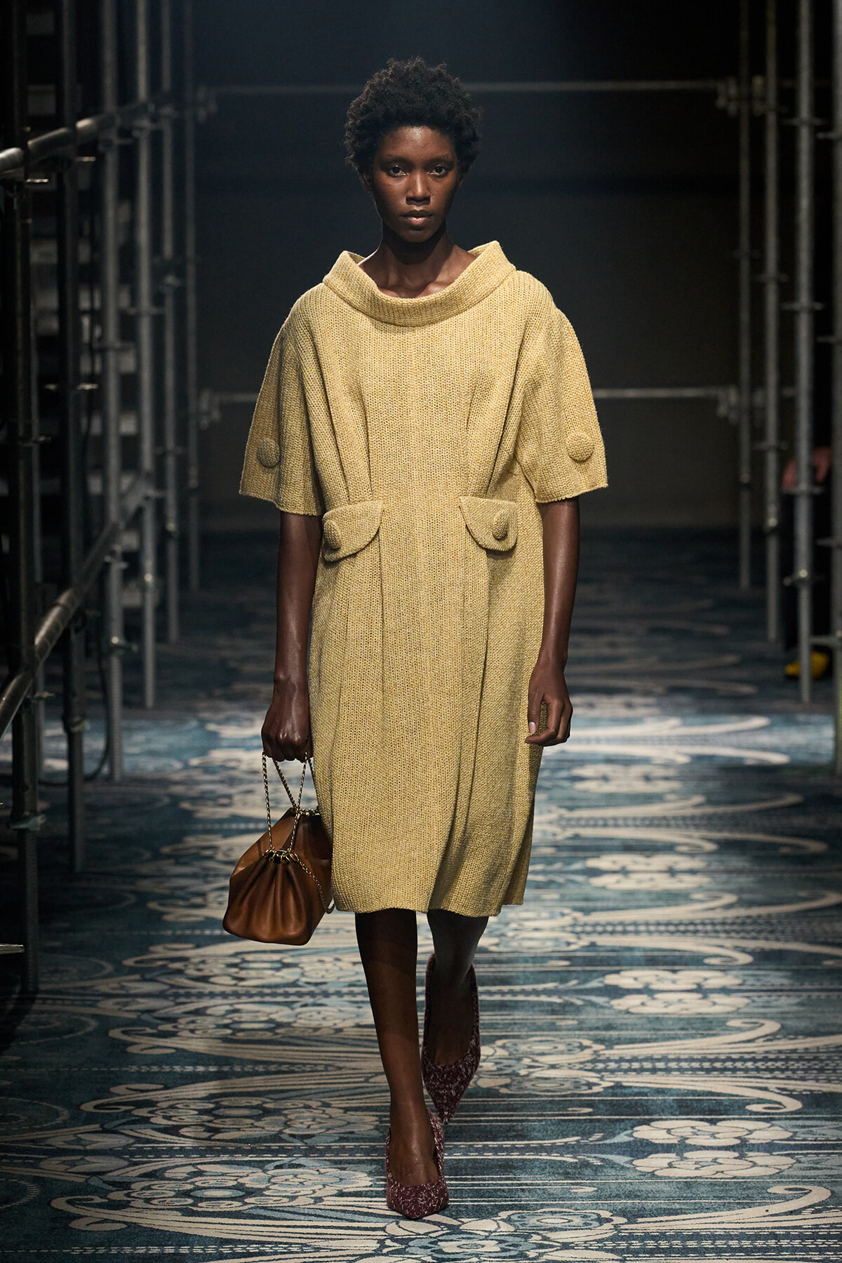 Black female model walking a runway in a beige mid-length knit dress with a wide cowl neck and large pocketed front, carrying a brown handbag.