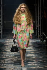 Model walking the runway in a bright, multicolor floral coat dress with long wavy hair, carrying a black handbag.