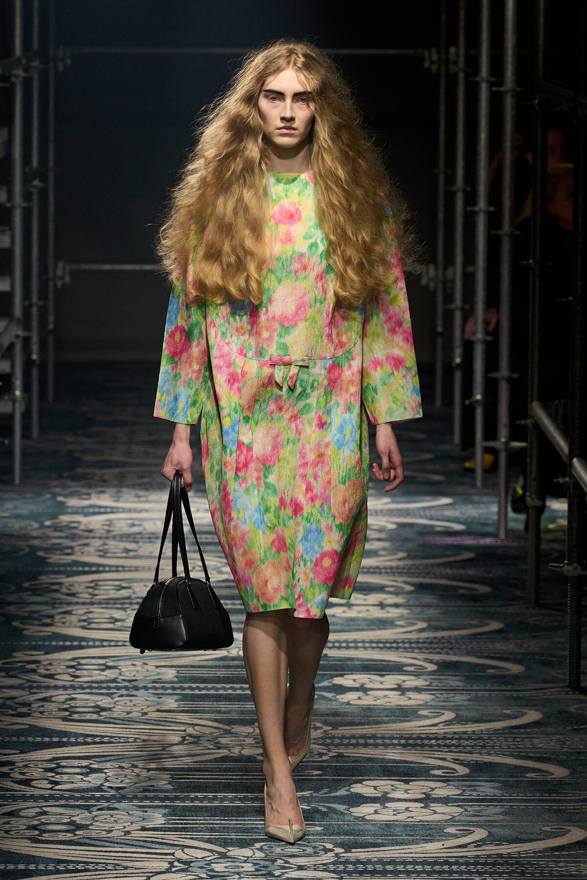 Model walking the runway in a bright, multicolor floral coat dress with long wavy hair, carrying a black handbag.