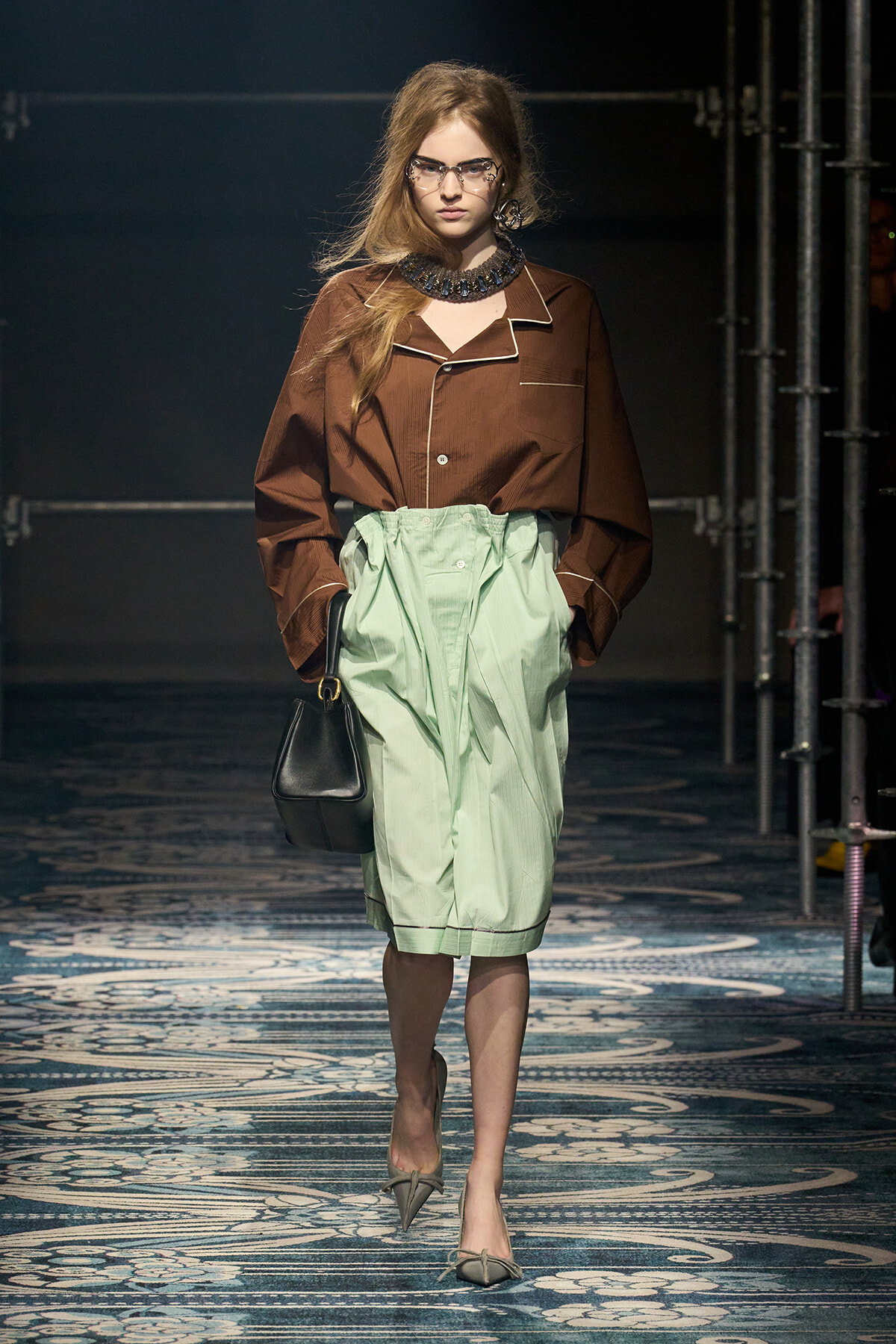Model walking runway in a brown button-down shirt with white piping and mint green high-waisted shorts, carrying a black handbag.