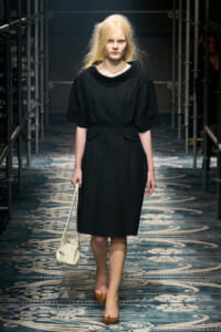 Model walking a runway in a black knee-length dress, holding a white handbag and wearing brown heels on a blue patterned carpet.