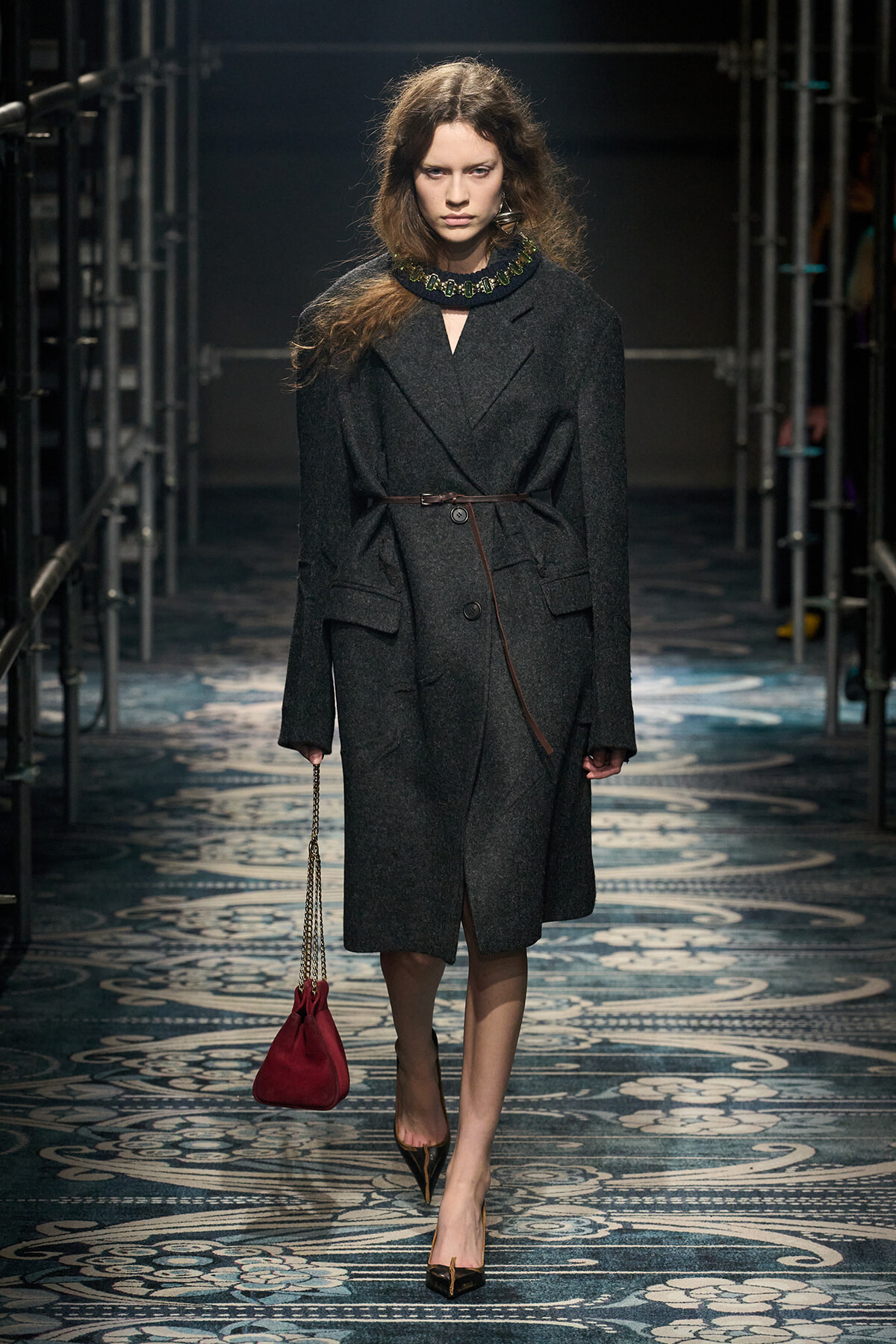 Model walks the runway in a dark gray wool coat cinched with a thin belt, paired with black heels and a red drawstring handbag on a chain.