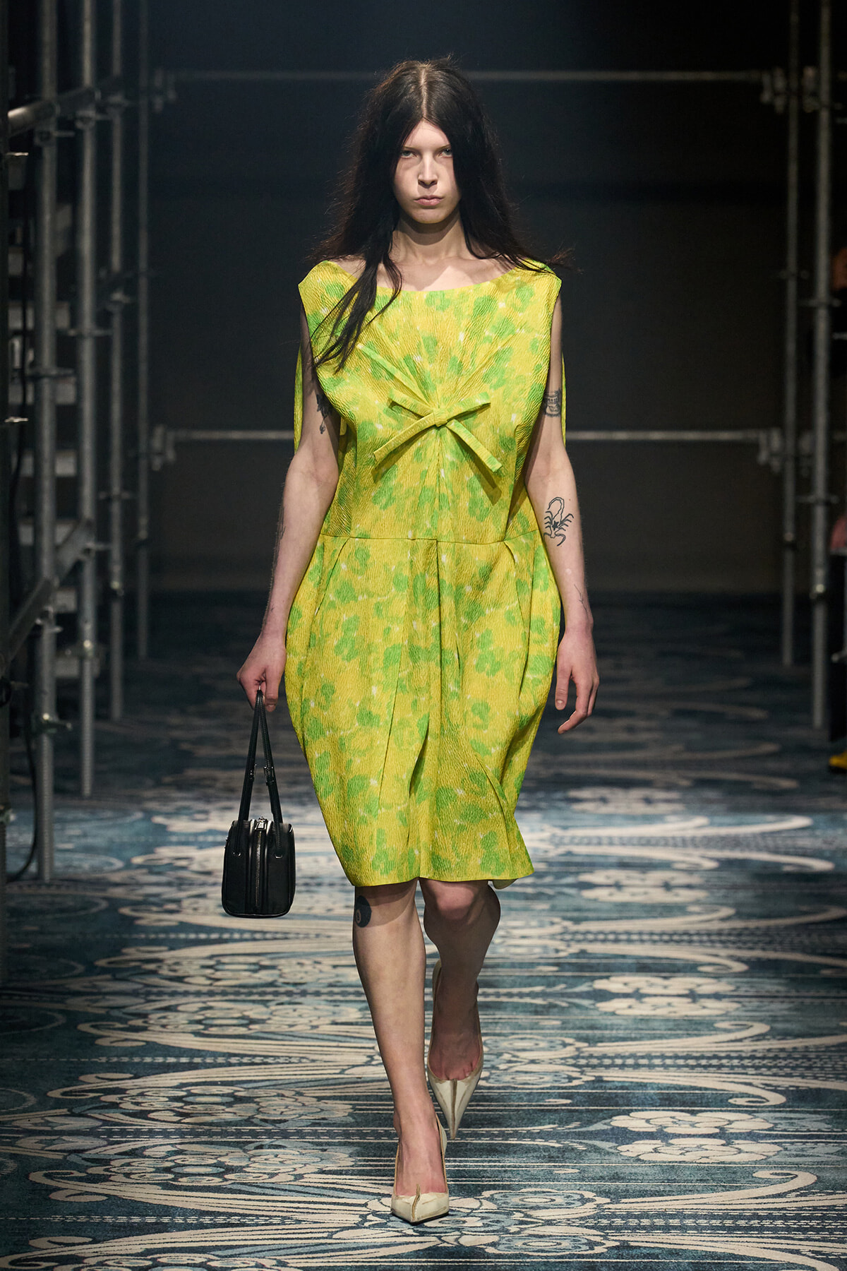 Model walking runway in a bright lime-green dress with a front bow, carrying a small black handbag and wearing beige heels.