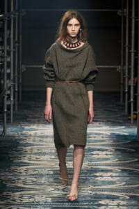 Model walks the runway in a gray knit midi dress belted at the waist, with a chunky pink necklace and beige sandals.