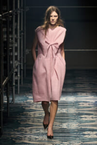 Model walks the runway wearing a pink knit sleeveless dress with a front bow and tied waist.