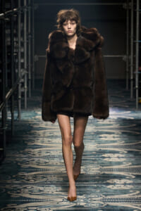 Model walks the runway in a dark brown oversized fur coat and tan heels on a patterned catwalk floor.