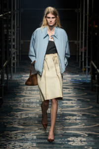 Model walking the runway wearing a light blue oversized jacket over a black top and beige knee-length skirt, with a brown shoulder bag and brown heels.