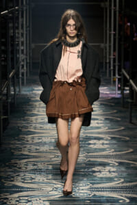 Model walks a fashion runway in a oversized black coat over a pale pink blouse and brown gathered mini skirt, wearing glasses and pointed heels.