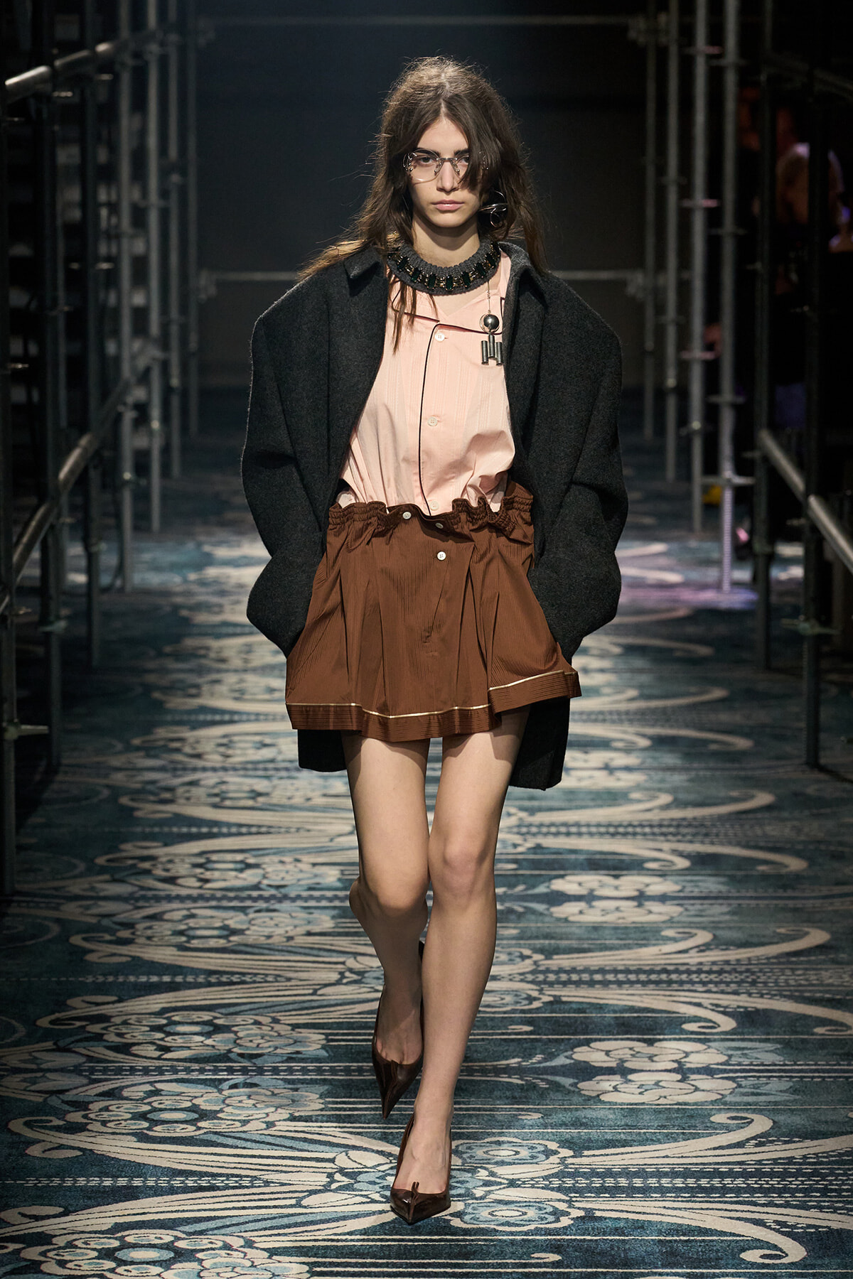 Model walks a fashion runway in a oversized black coat over a pale pink blouse and brown gathered mini skirt, wearing glasses and pointed heels.