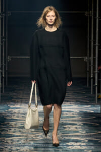 Model walking a runway in a knee-length black dress, carrying a white handbag and wearing pointed brown heels.