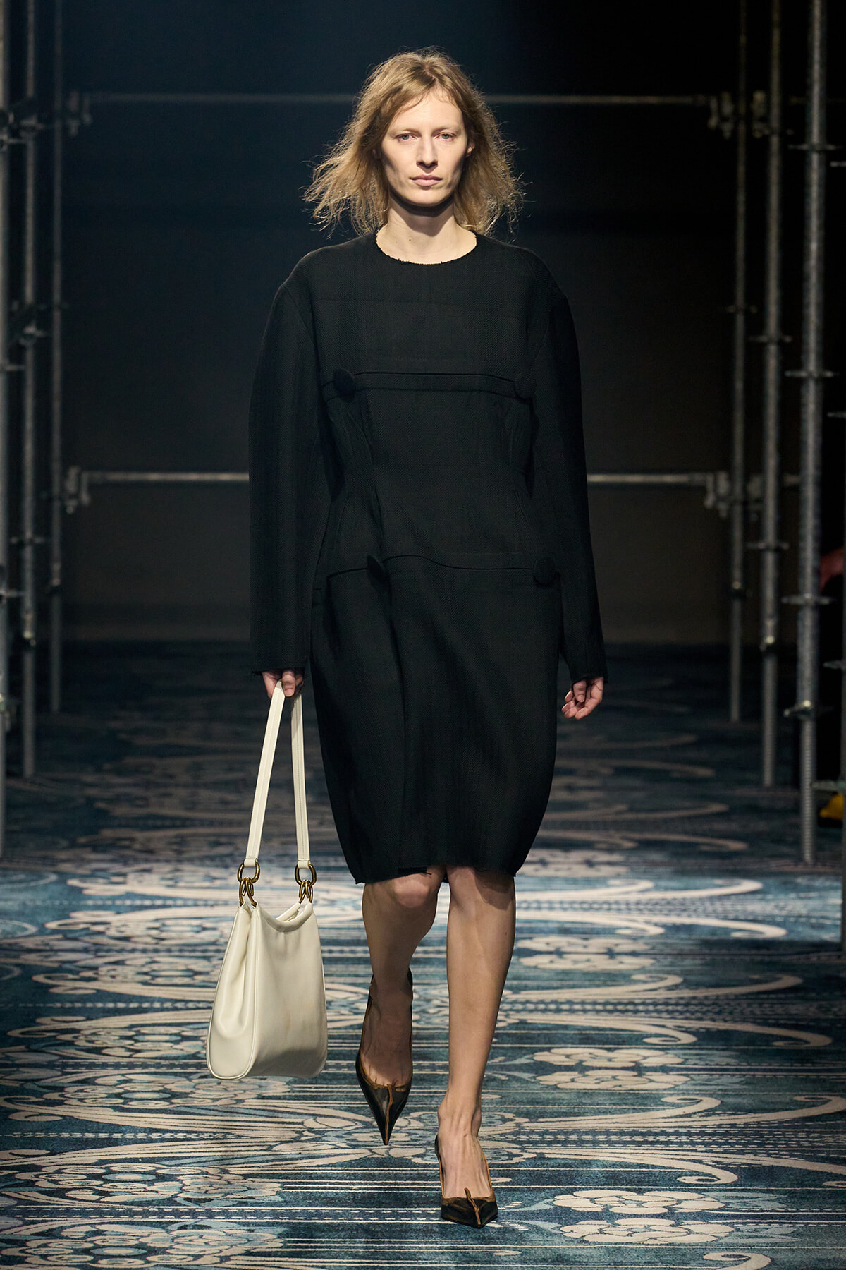 Model walking a runway in a knee-length black dress, carrying a white handbag and wearing pointed brown heels.