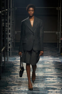 Model walks the runway in a dark grey, oversized blazer with a matching skirt, carrying a small black handbag.