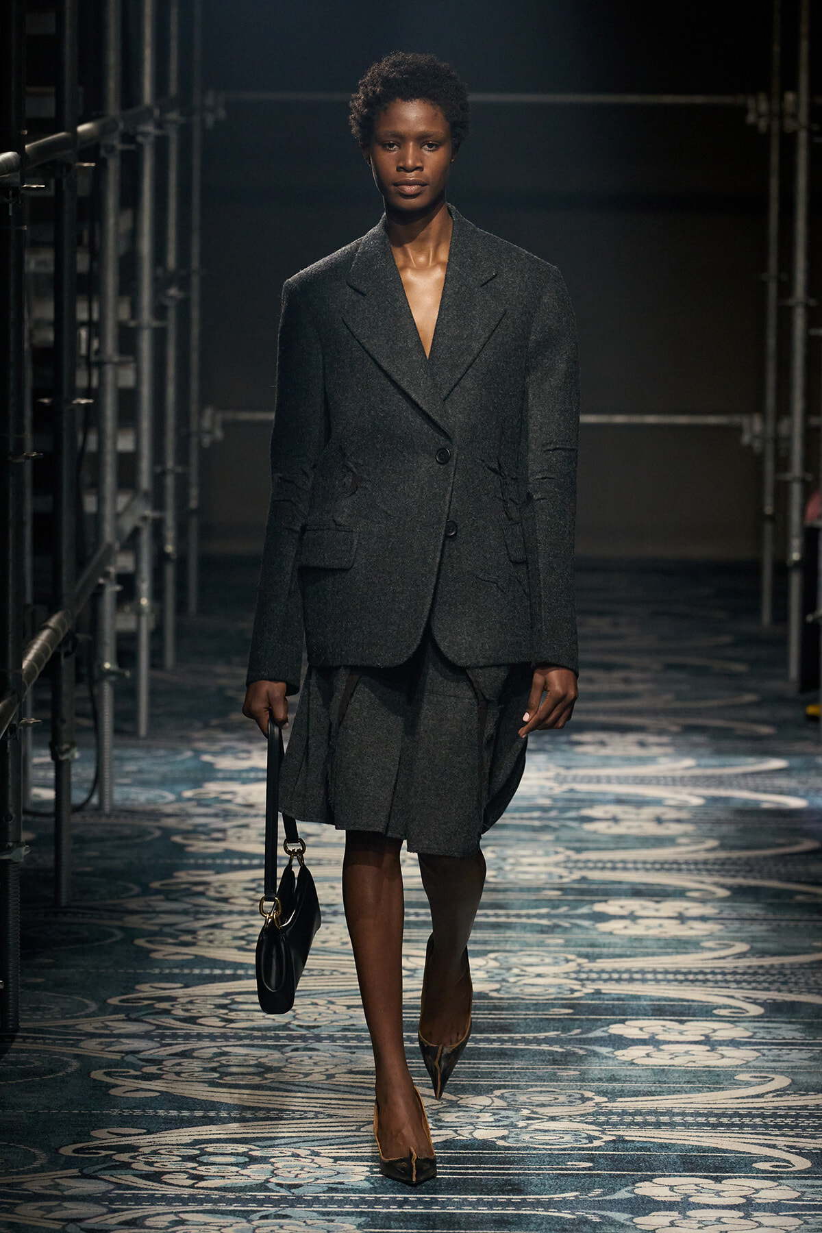 Model walks the runway in a dark grey, oversized blazer with a matching skirt, carrying a small black handbag.