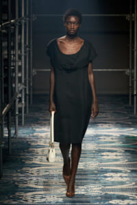 Model walking a fashion runway in a black knee-length dress, holding a white handbag and wearing brown heels on a patterned catwalk backdrop.