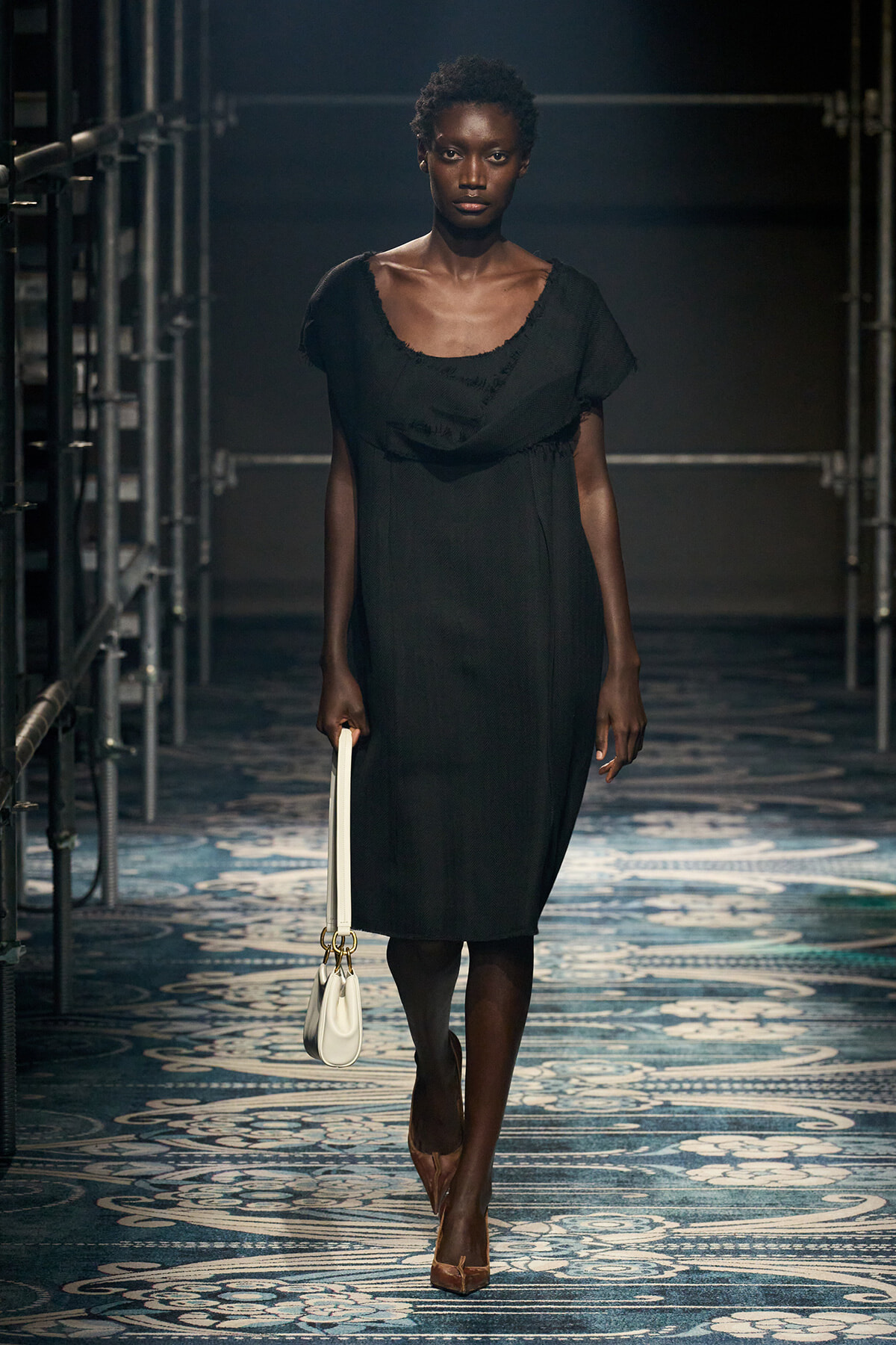 Model walking a fashion runway in a black knee-length dress, holding a white handbag and wearing brown heels on a patterned catwalk backdrop.