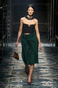 Model walking a fashion runway in a black strapless top and dark green wrapped midi skirt, accessorized with a chunky beaded necklace and a small brown handbag, wearing maroon glitter heels.