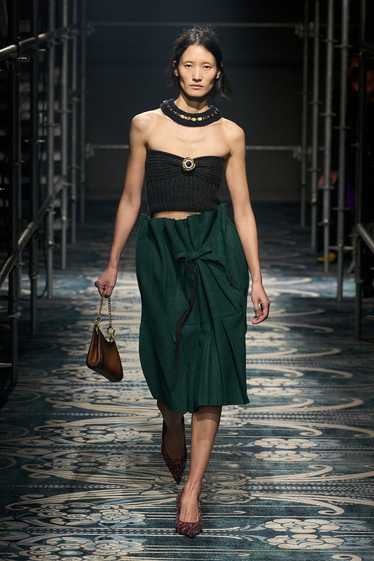 Model walking a fashion runway in a black strapless top and dark green wrapped midi skirt, accessorized with a chunky beaded necklace and a small brown handbag, wearing maroon glitter heels.