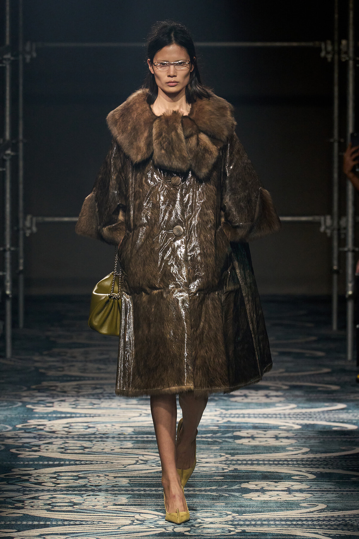 Model wears a long brown fur coat with a large fur collar, sunglasses, and yellow heels on a runway, carrying a mustard handbag