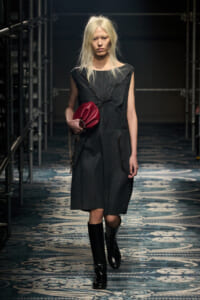 Model walking a fashion runway in a dark gray knee-length dress with a tied waist, black knee-high boots, and a red clutch.