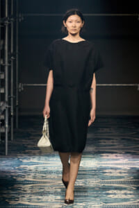 Model walking the runway in a loose black dress, carrying a small white handbag, backstage with scaffolding visible.