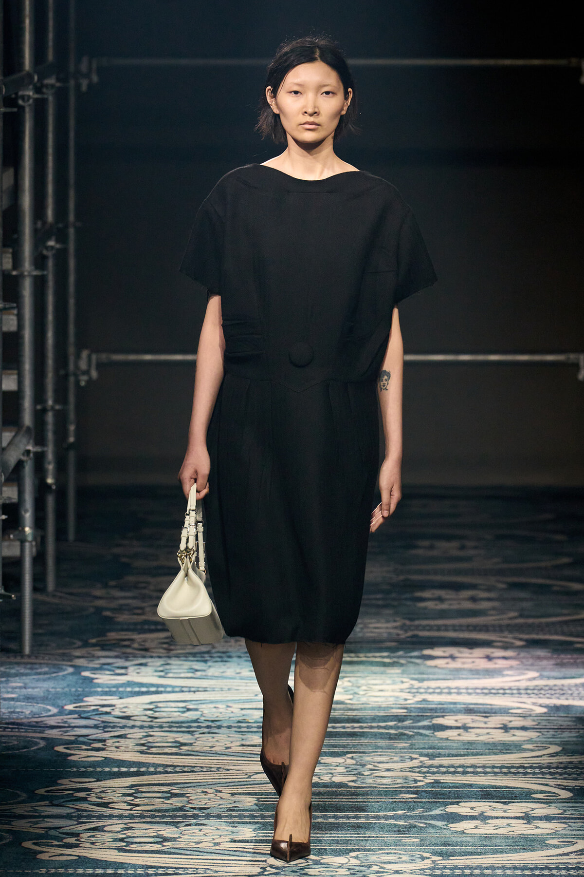 Model walking the runway in a loose black dress, carrying a small white handbag, backstage with scaffolding visible.