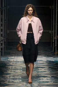 Model walks a fashion show in a light pink oversized jacket over a brown crop top and black skirt, carrying a brown handbag.