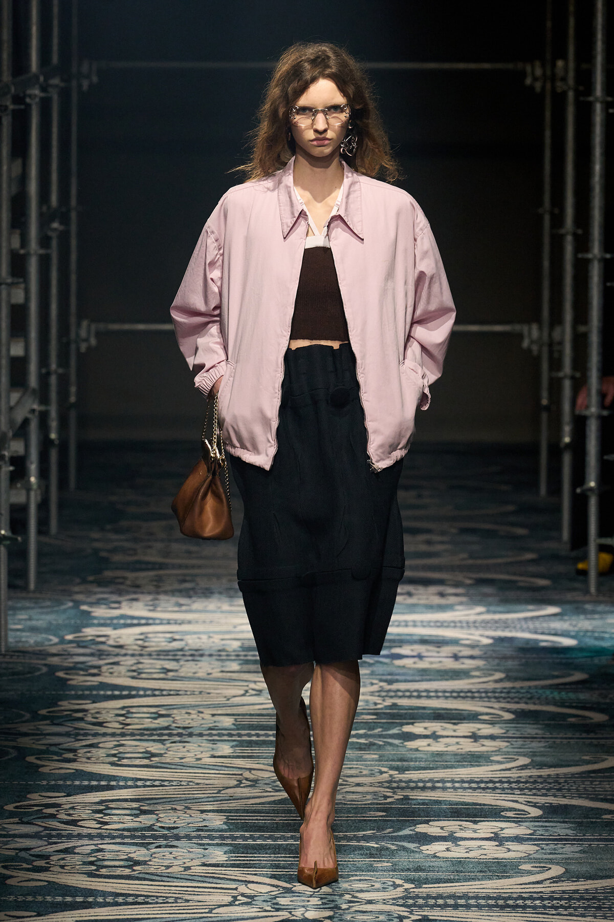 Model walks a fashion show in a light pink oversized jacket over a brown crop top and black skirt, carrying a brown handbag.