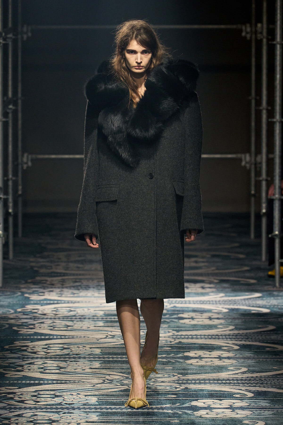 Female model on a runway wearing a long dark wool coat with an oversized black fur collar.