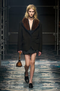Model walking the runway in a dark blazer with a large brown fur collar, black skirt, and black loafers, carrying a small brown handbag.