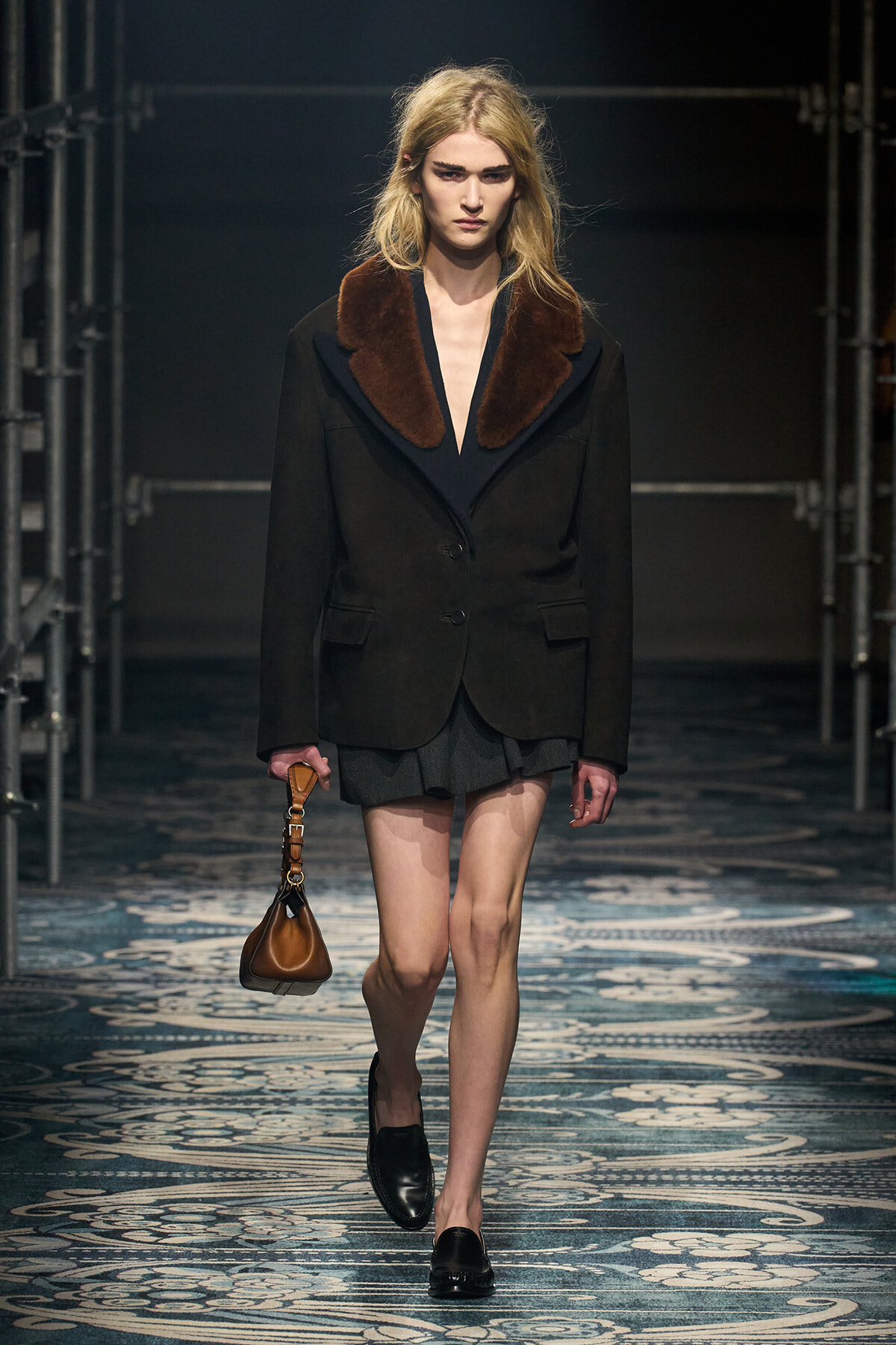 Model walking the runway in a dark blazer with a large brown fur collar, black skirt, and black loafers, carrying a small brown handbag.