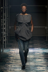 Model walks a fashion runway wearing a gray twisted-front top over jeans in a high-fashion look, backstage with metal scaffolding nearby.