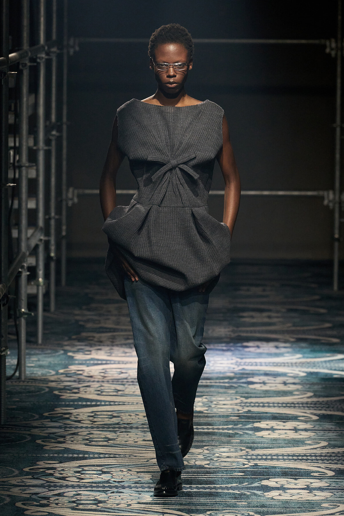 Model walks a fashion runway wearing a gray twisted-front top over jeans in a high-fashion look, backstage with metal scaffolding nearby.