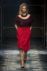 Model walking a runway in a burgundy off-the-shoulder top and bright red midi skirt with a bow belt, paired with a chunky black necklace.