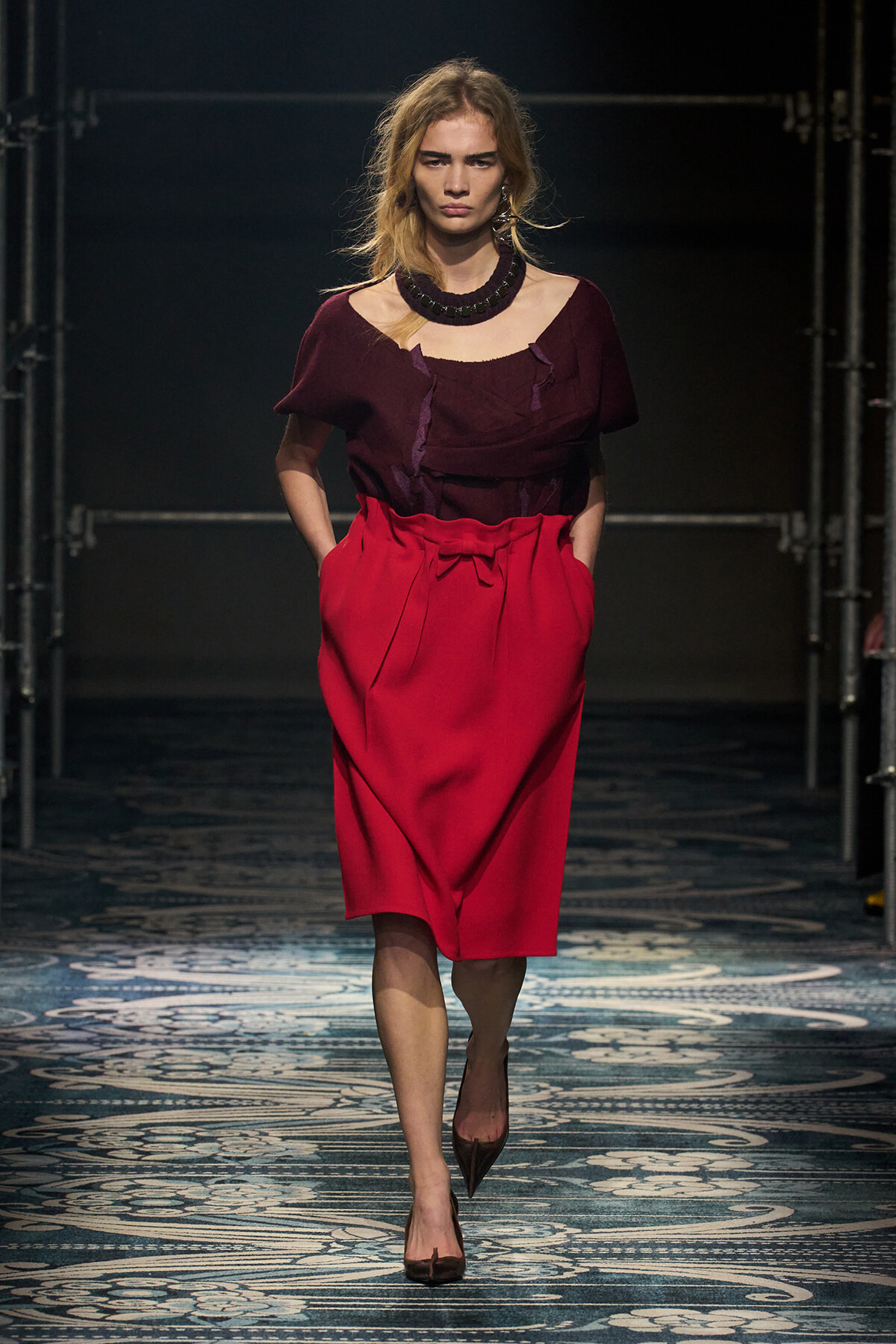 Model walking a runway in a burgundy off-the-shoulder top and bright red midi skirt with a bow belt, paired with a chunky black necklace.