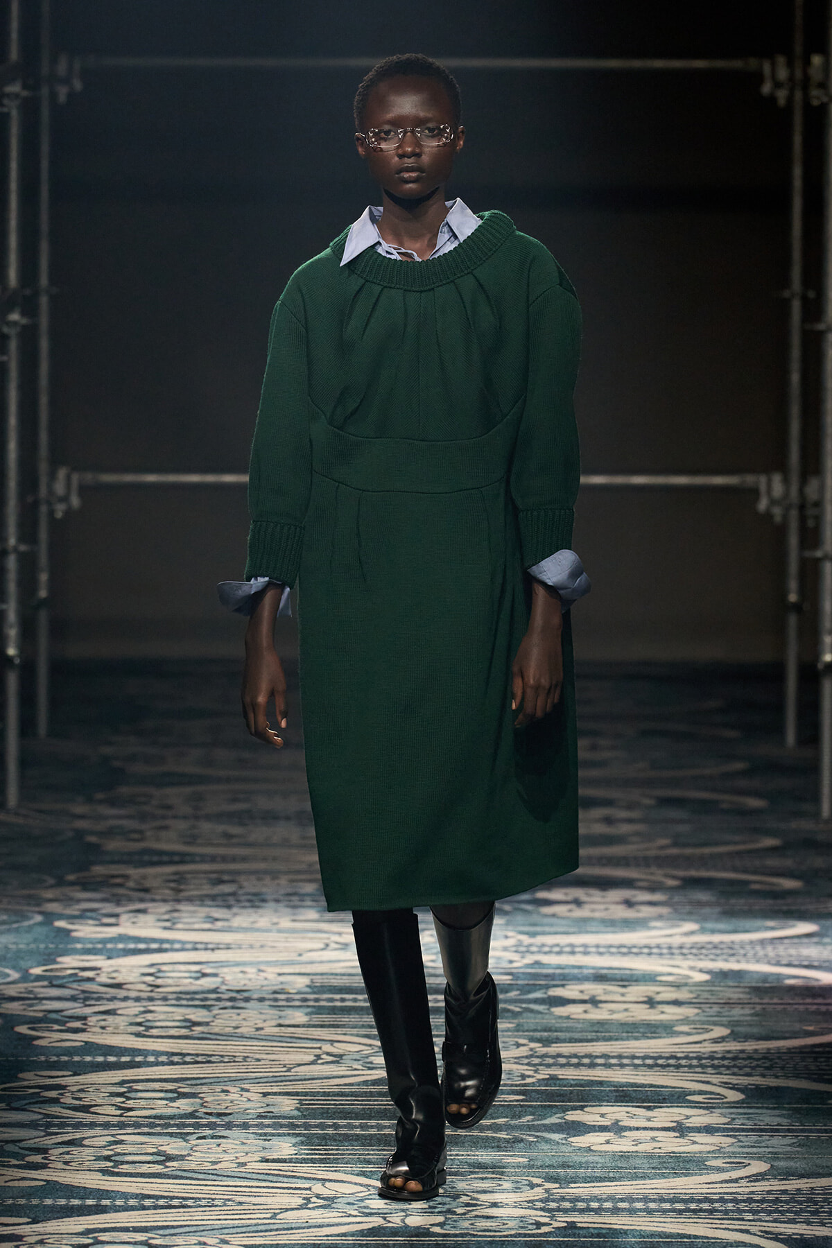 Model walking the runway in a dark emerald-green knit dress layered over a blue shirt, with clear glasses and black knee-high boots