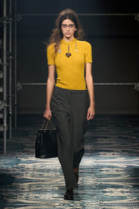 Model walking a runway in a mustard yellow short-sleeve knit top, gray tailored trousers, and a black handbag with chain straps.