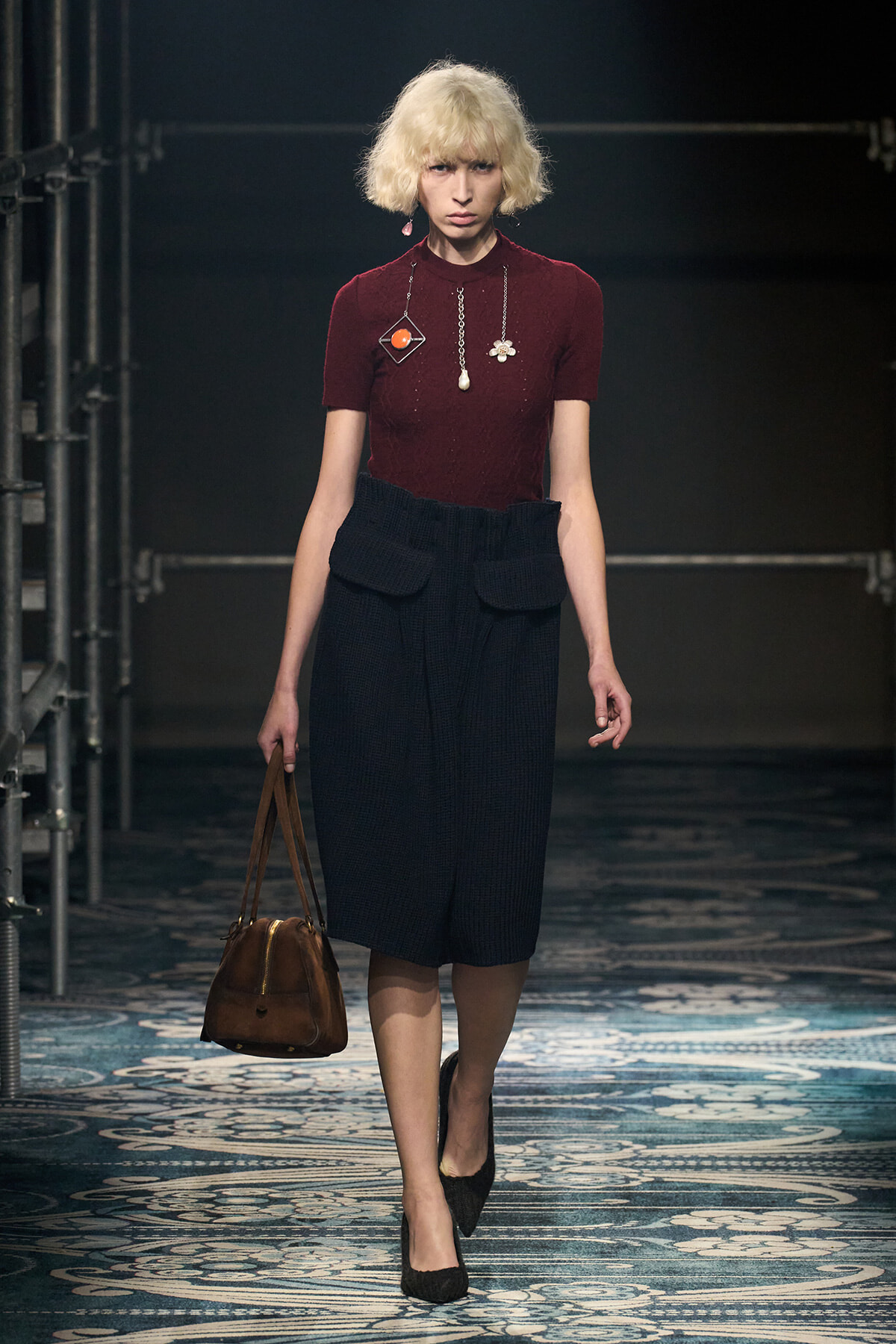 Model walking a runway in a burgundy knit tee and navy high-waisted skirt with large pockets, carrying a brown handbag.