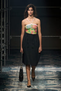 Model walking on a runway in a strapless colorful top with a black midi skirt, carrying a black handbag.