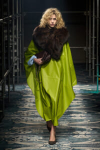 Model walking the runway in a lime-green cape coat with a large dark fur stole, holding a brown clutch and chain accessory; blue shirt cuffs peek out, curly blonde hair.