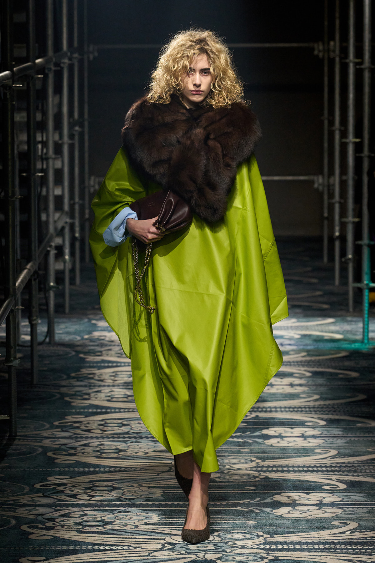 Model walking the runway in a lime-green cape coat with a large dark fur stole, holding a brown clutch and chain accessory; blue shirt cuffs peek out, curly blonde hair.