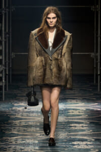 Model walks a fashion runway wearing a brown fur coat with dark lapels, white blouse, black loafers, and a small black handbag.
