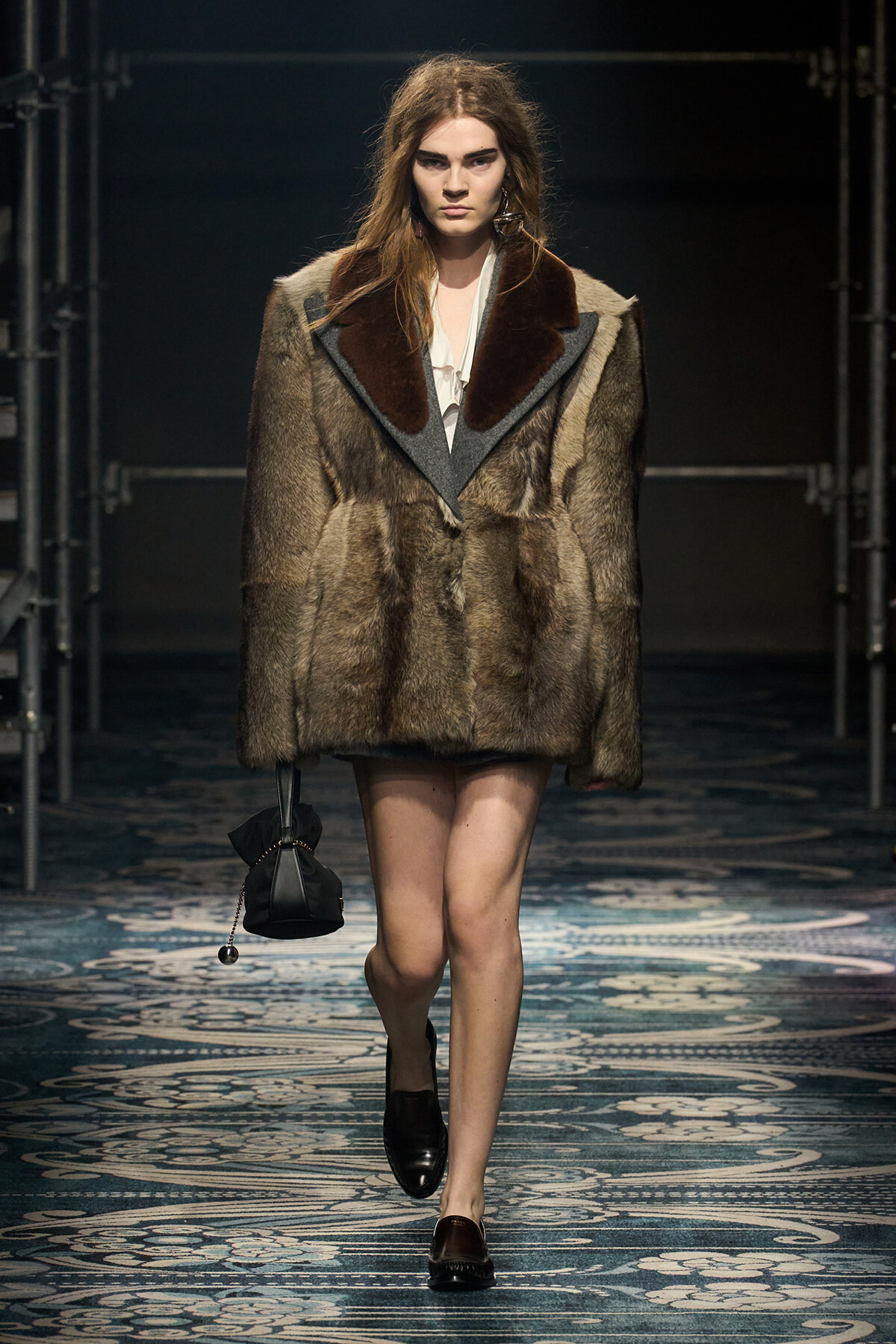 Model walks a fashion runway wearing a brown fur coat with dark lapels, white blouse, black loafers, and a small black handbag.