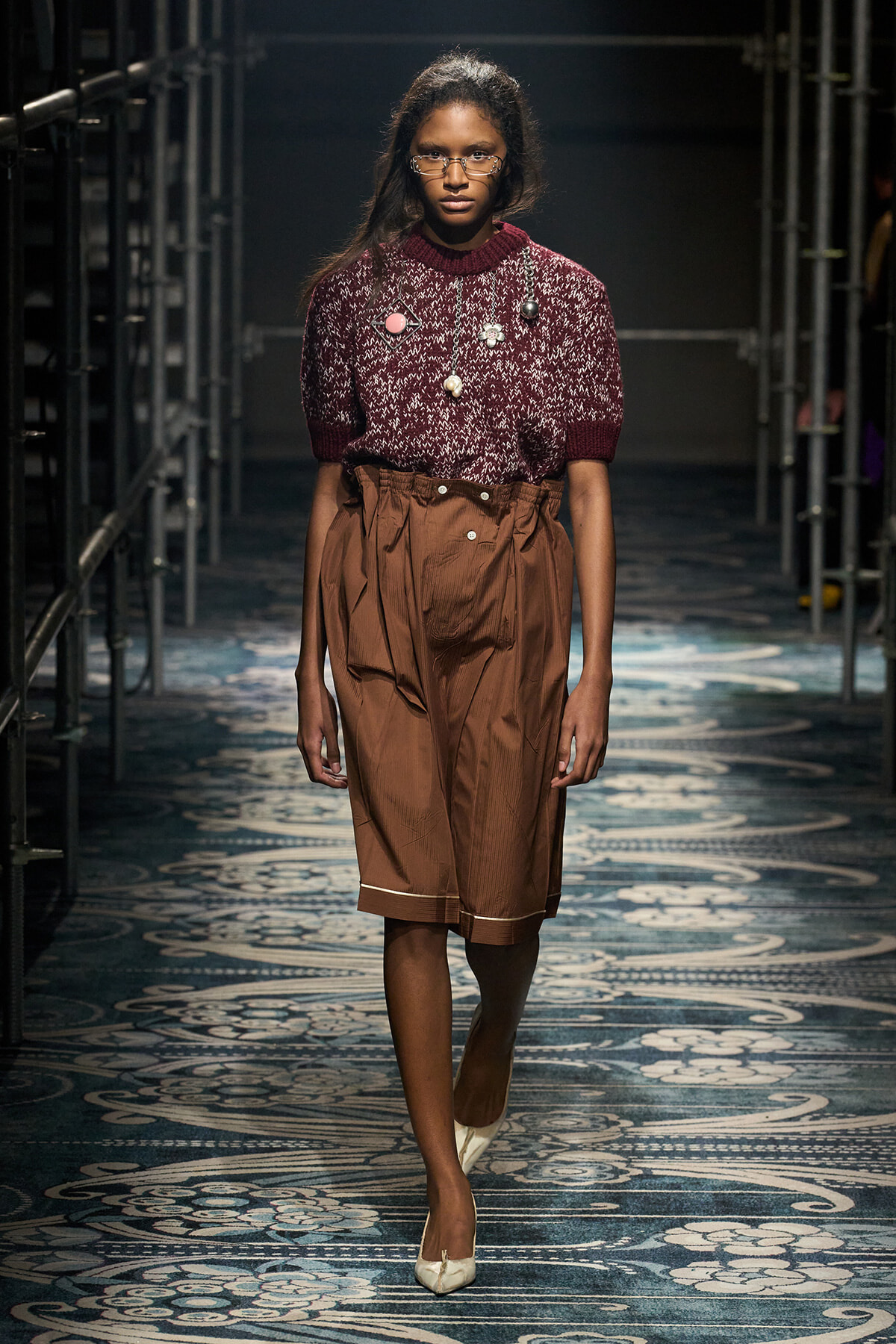 Female model walking a runway in a burgundy knit sweater and high-waisted brown shorts with beige heels.