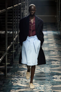 Model walks a runway in a long dark overcoat, burgundy shirt with yellow piping, and oversized light-blue shorts, beige sneakers.