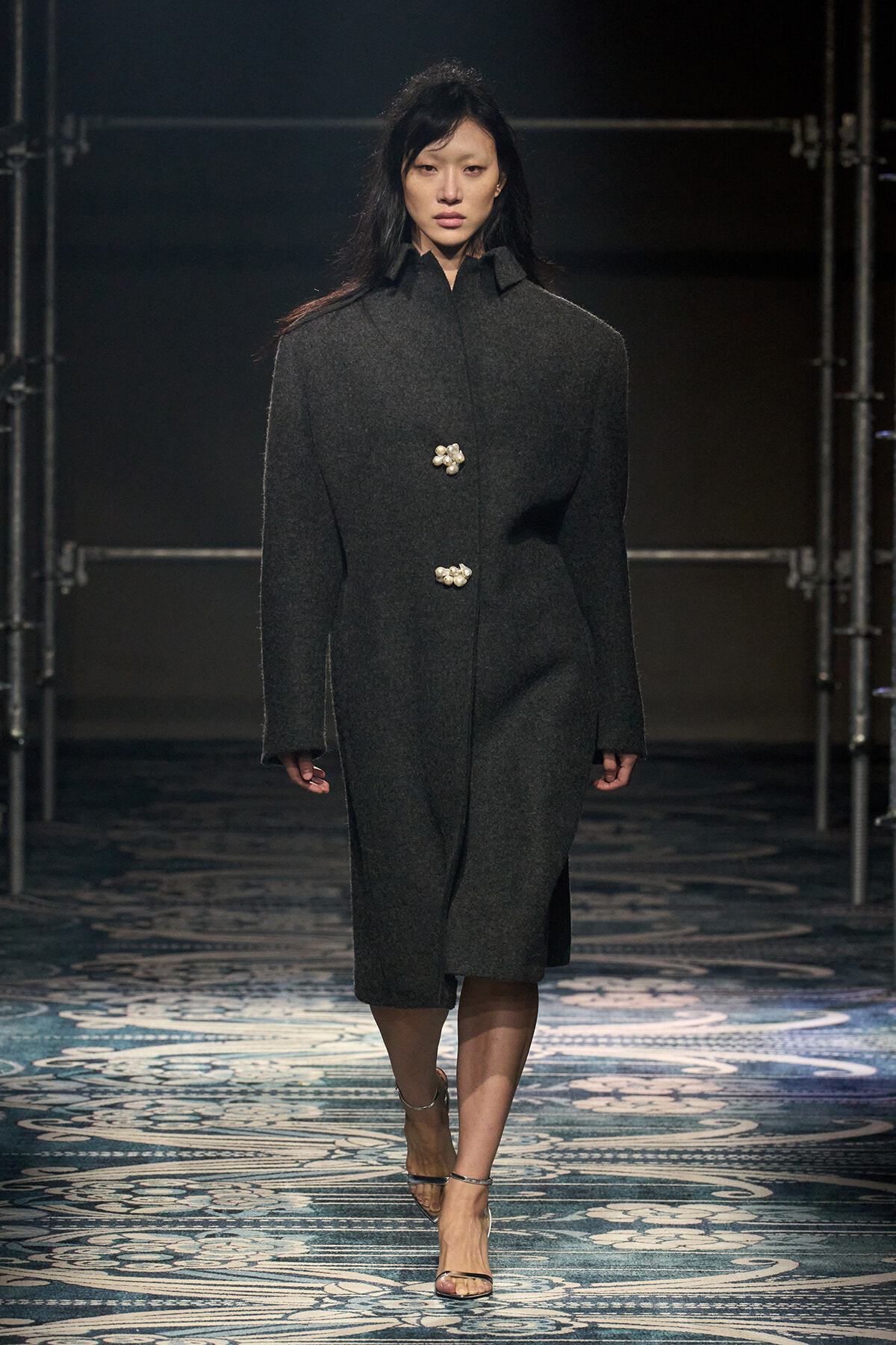 Model walking a fashion runway in a dark gray knee-length wool coat with two pearl-like buttons on the front, against a scaffolded backdrop and patterned floor.