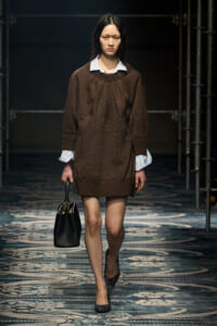 Model walking a runway in a brown knit dress with white shirt cuffs, carrying a black handbag and wearing dark, textured heels.
