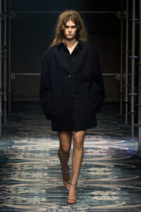 Female model walking a fashion runway in a black oversized coat with large buttons, hands in pockets on a patterned catwalk.